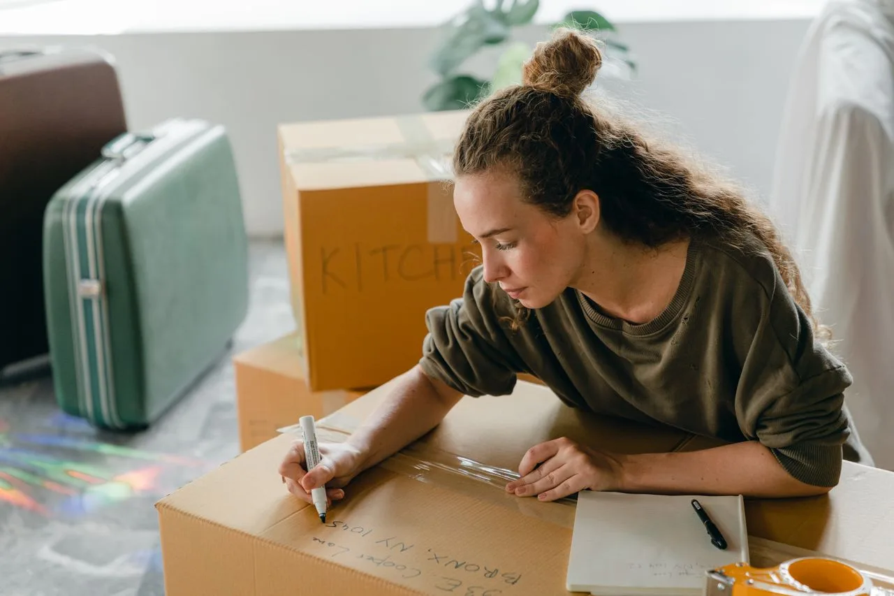 our-journey-03 Top view of thoughtful lady in casual wear writing with marker on cardboard box while sitting near suitcases and pile of boxes in light living room