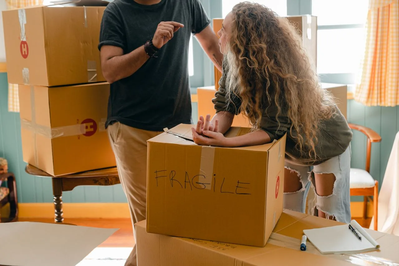who-we-are Crop faceless man in wristwatch talking to girlfriend in fashion jeans with wavy hair while standing near box with FRAGILE note after moving into new house